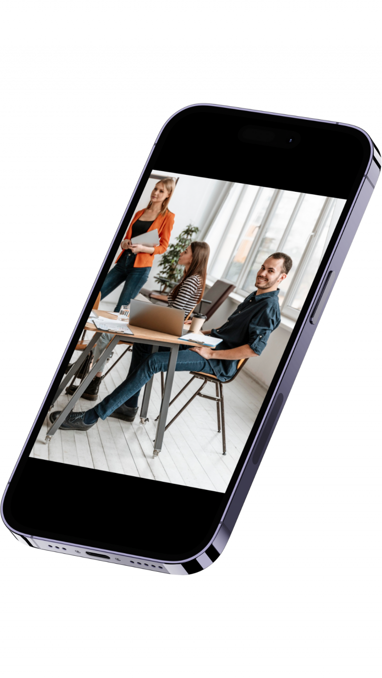 Group of colleagues in a bright office around a table, one woman presenting with a laptop while two others listen and work.